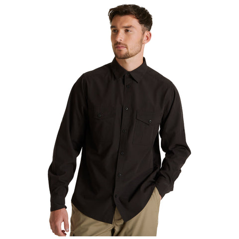 A man wearing a black long-sleeve shirt with two pockets stands in a neutral setting looking slightly to the side with a relaxed expression and one hand in his pocket.