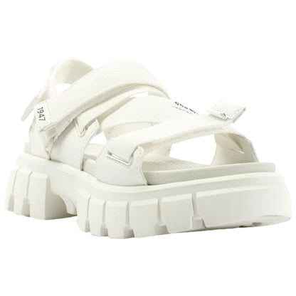 White sandals feature thick straps and a chunky sole. They rest on a flat surface, showcasing a modern, casual design suitable for warm weather or relaxed settings.