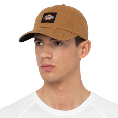A young man is wearing a brown baseball cap with a black logo patch on the front while looking slightly to the side against a plain white background.