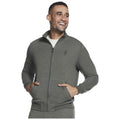 A man is smiling while wearing a green zip-up jacket with a high collar and front pockets in a neutral background. His hands are placed in the pockets.