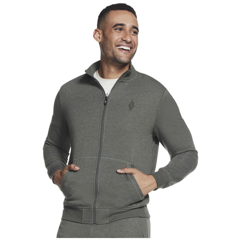 A man is smiling while wearing a green zip-up jacket with a high collar and front pockets in a neutral background. His hands are placed in the pockets.