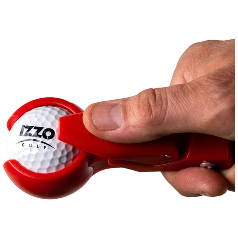 A hand grips a red golf ball cleaner tool as it presses against a white golf ball displaying the logo "IZZO GOLF" in a bright, uncluttered setting.