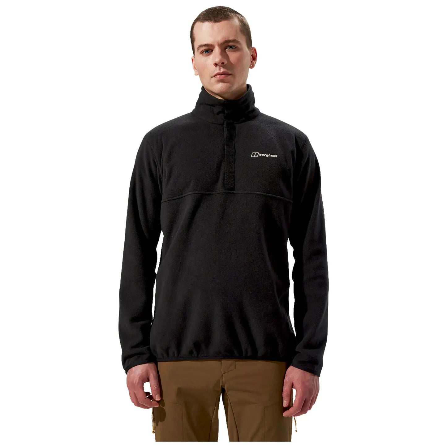 A man stands upright wearing a black fleece pullover with a high collar and a logo on the chest paired with brown pants in a neutral background.