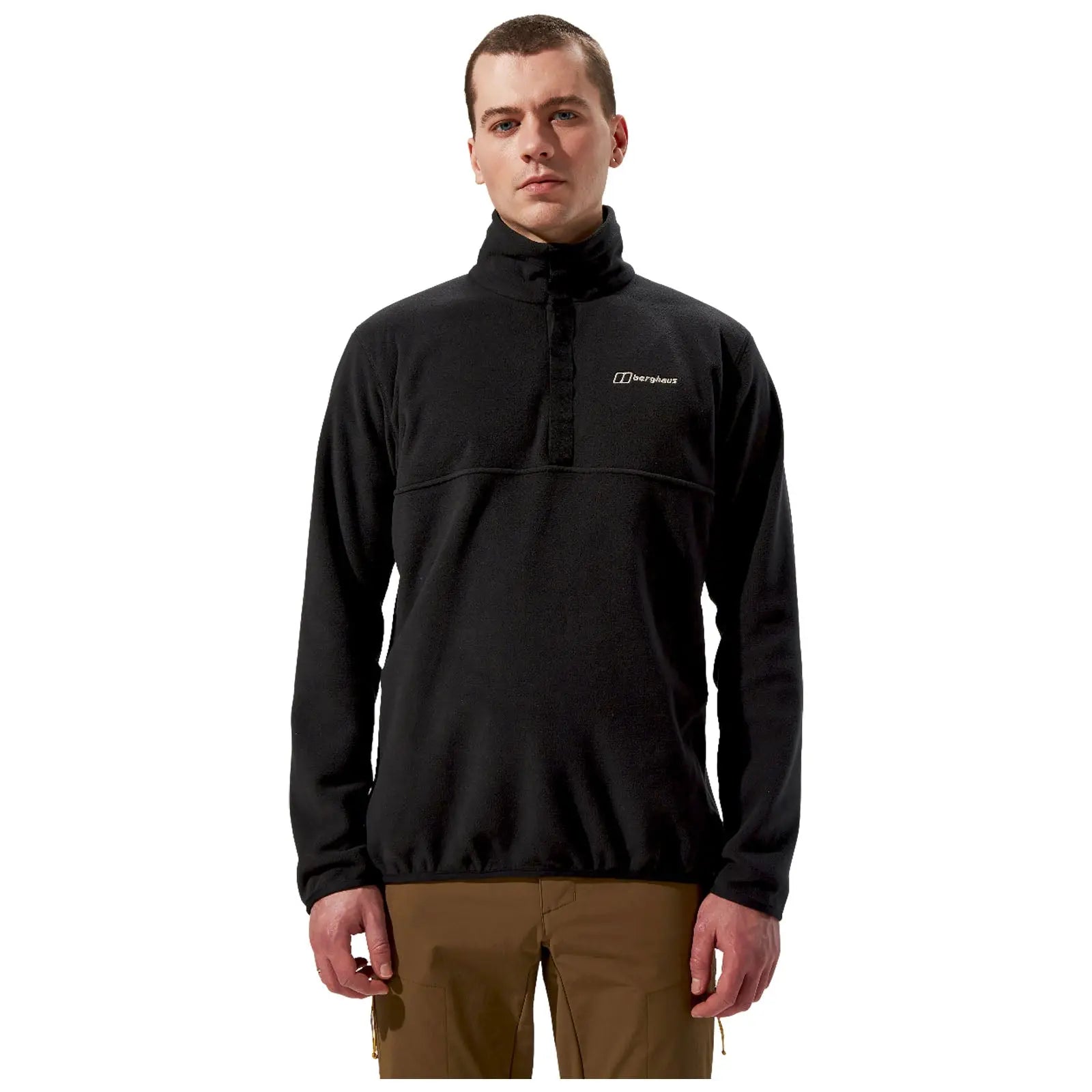 A man stands upright wearing a black fleece pullover with a high collar and a logo on the chest paired with brown pants in a neutral background.