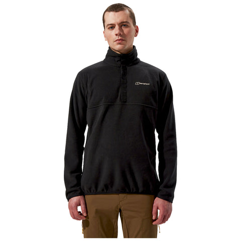A man stands upright wearing a black fleece pullover with a high collar and a logo on the chest paired with brown pants in a neutral background.
