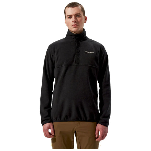 A man stands upright wearing a black fleece pullover with a high collar and a logo on the chest paired with brown pants in a neutral background.