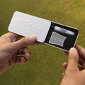 A hand holds a white box with "Titleist" embossed on it containing a marker and a comb sitting on black foam, set against a green grass background.