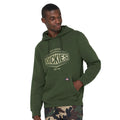 A man is wearing a green hoodie with the brand name Dickies printed on it while standing with one hand in the pocket and a slight smile against a plain background.