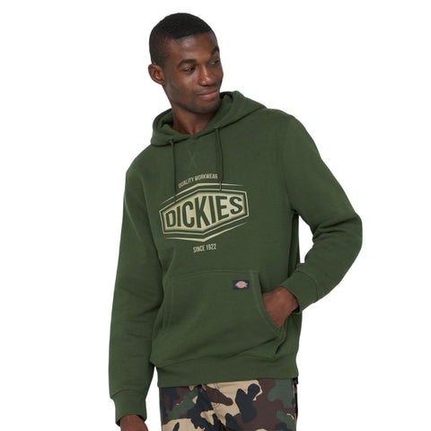 A man is wearing a green hoodie with the brand name Dickies printed on it while standing with one hand in the pocket and a slight smile against a plain background.