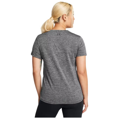 A woman stands facing away wearing a short-sleeved gray athletic shirt paired with black leggings her hair is styled in a ponytail and she has visible tattoos on her arms.
