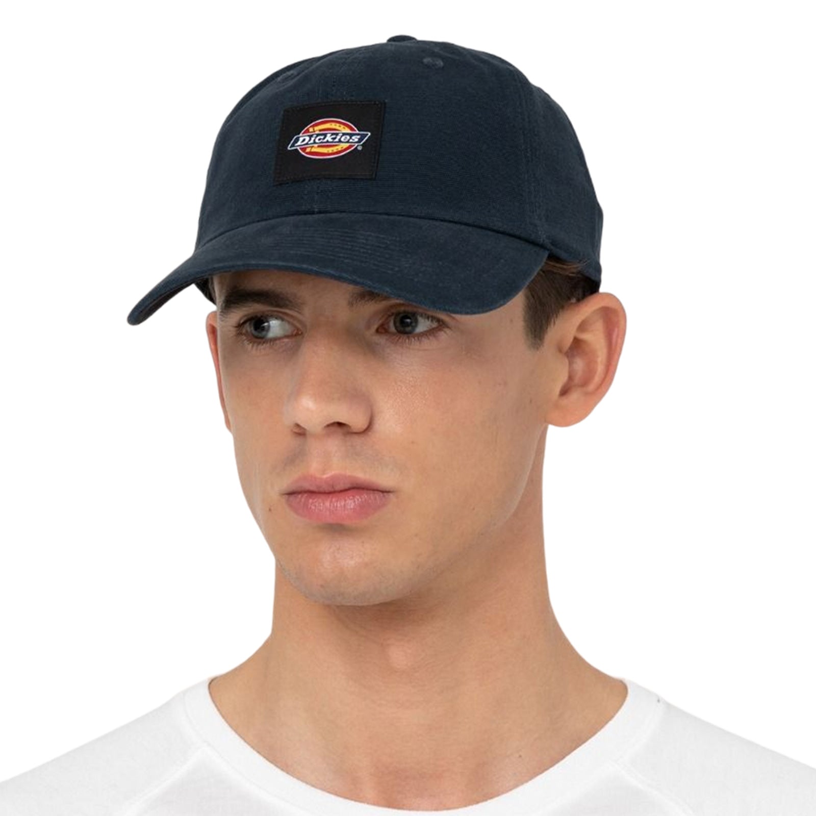 A man wears a dark cap with a logo while looking slightly to the side in a neutral expression against a plain background, dressed in a white shirt.