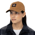 A person wears a brown cap featuring a logo while looking slightly to the side. They are dressed in a black and gray plaid shirt, standing against a plain white background.