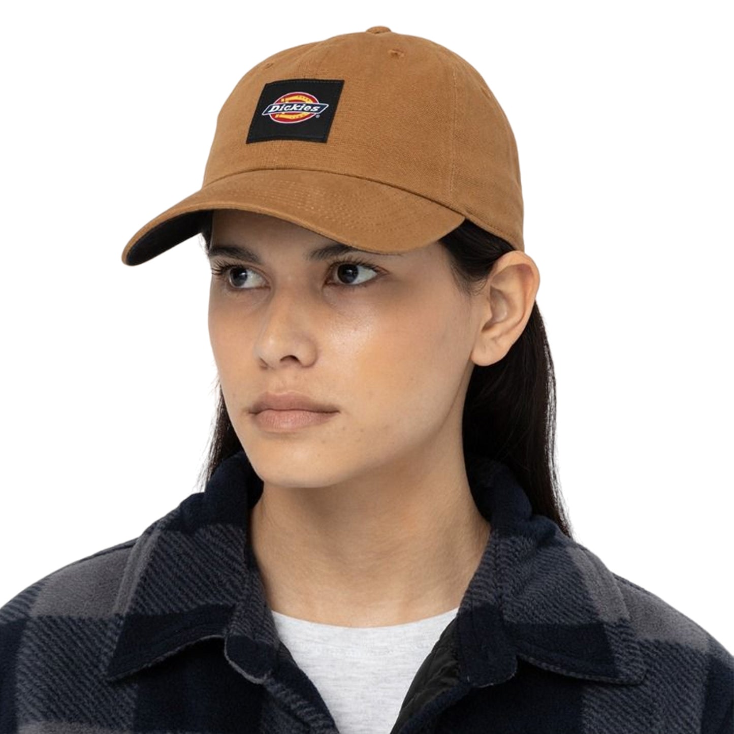 A person wears a brown cap featuring a logo while looking slightly to the side. They are dressed in a black and gray plaid shirt, standing against a plain white background.