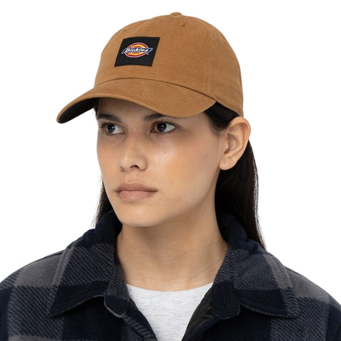 A person wears a brown cap featuring a logo while looking slightly to the side. They are dressed in a black and gray plaid shirt, standing against a plain white background.