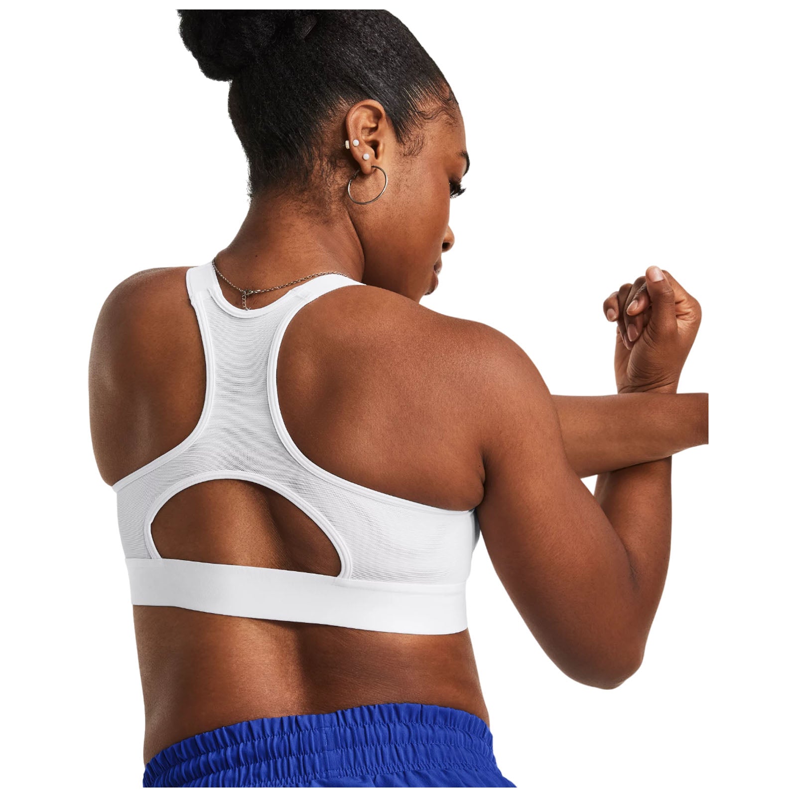 A woman in a white athletic tank top adjusts her arm while facing away from the viewer. She has braided hair and is wearing small earrings in a bright setting.