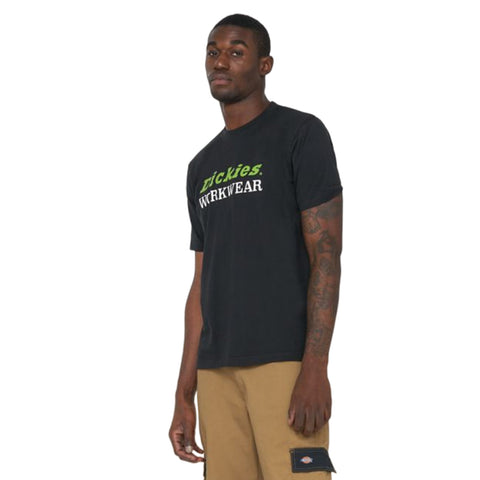 A young man stands confidently wearing a black t-shirt with the text "trickies. WORKWEAR" in green and white. He pairs it with tan cargo shorts.