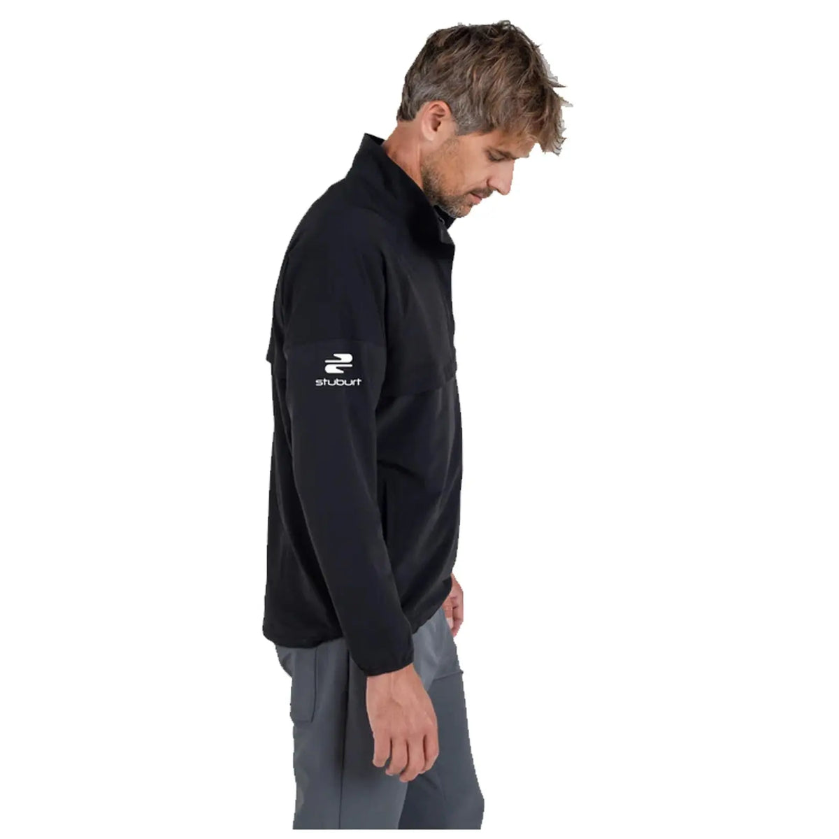 A man is standing sideways wearing a black jacket with a high collar and gray pants. He appears to be looking down, with a focused expression. The jacket features a logo on the sleeve.
