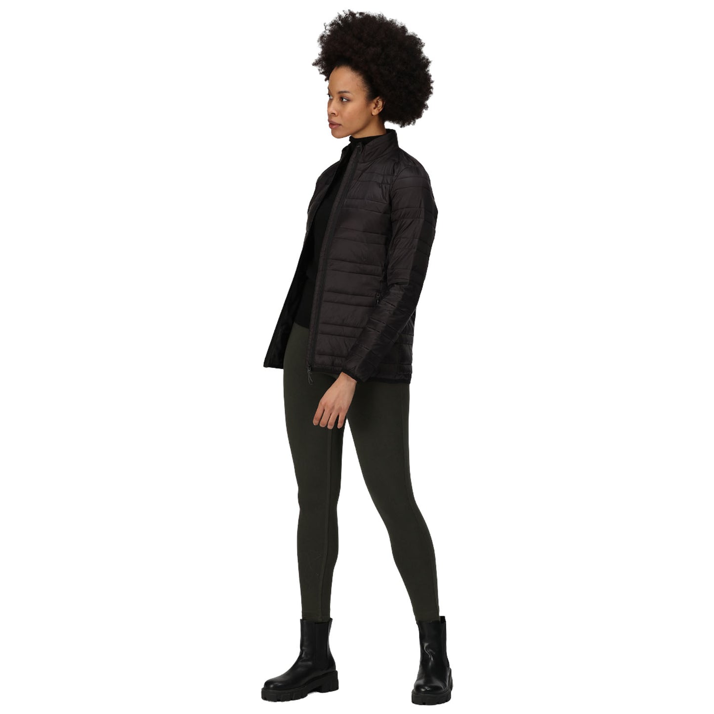 A person stands in a black padded jacket with the front unzipped wearing dark leggings and black boots in a neutral space that highlights their outfit and posture.