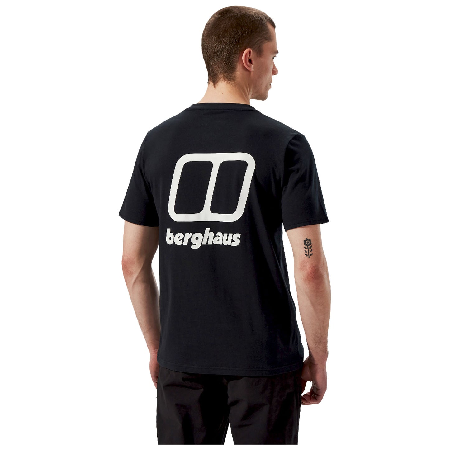 A person stands wearing a black t-shirt featuring a large white logo and the word "berghaus" on the back in a plain background. The individual has a tattoo on their arm.