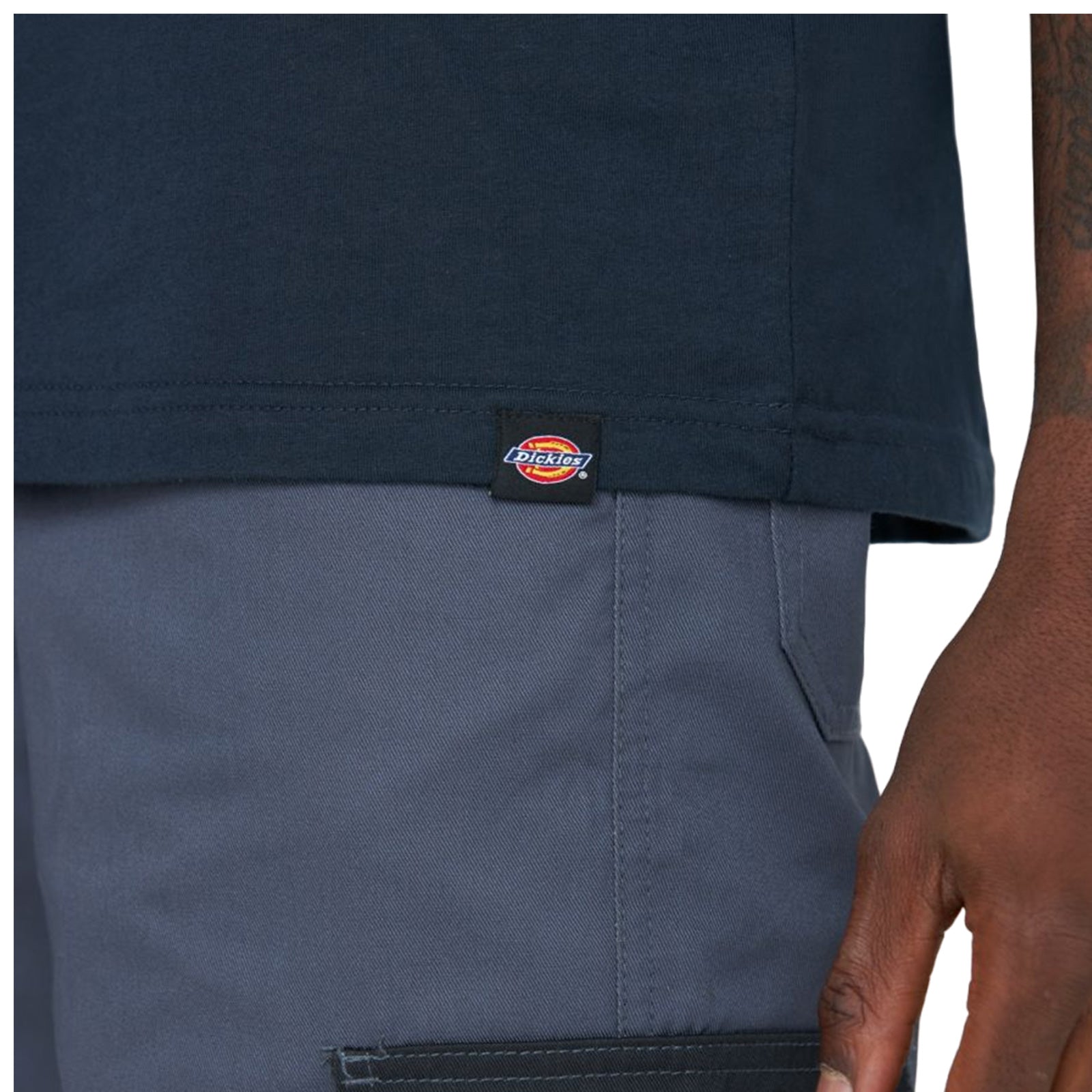 A person's hand holds the edge of a navy blue shirt above gray pants featuring a visible brand label on the shirt's hem and the pants' pocket area.