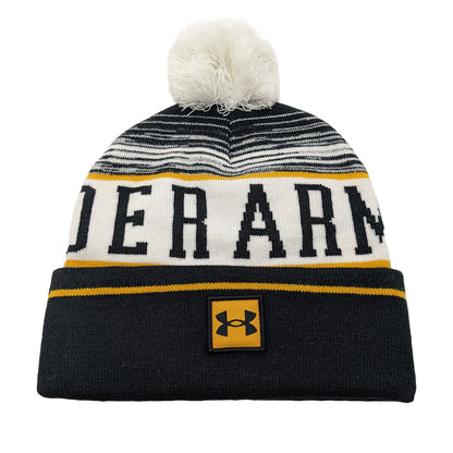 A knitted beanie with a white and black striped pattern features bold yellow and black letters spelling "UNDER ARMOUR" on the body and a logo patch sewn on the front. A fluffy pom-pom sits atop the hat.