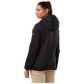 A woman stands facing slightly away wearing a black quilted jacket with a hood and light-colored pants in a neutral background showcasing a stylish outdoor look.