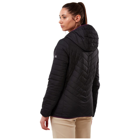 A woman stands facing slightly away wearing a black quilted jacket with a hood and light-colored pants in a neutral background showcasing a stylish outdoor look.