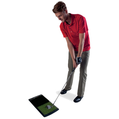 A man in a red polo shirt is preparing to swing a golf club while standing on a practice mat with a green turf surface in a plain background setting.