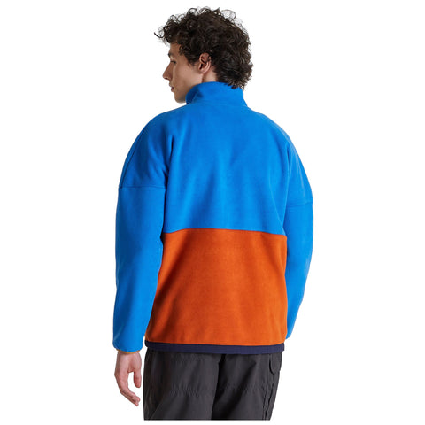A man wearing a two-toned fleece jacket stands with his back to the viewer showcasing a blue upper section and an orange lower section in a neutral background