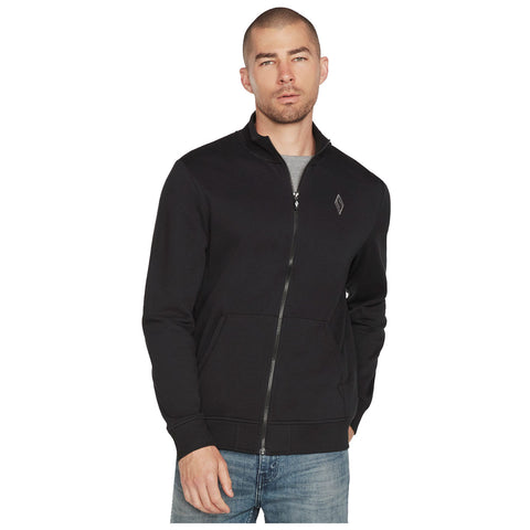 A man is standing while wearing a black zip-up sweatshirt over a gray shirt and blue jeans with his hands in his pockets against a plain white background.