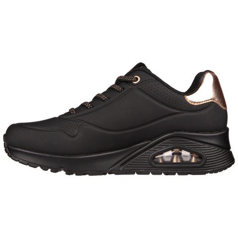 A black athletic shoe features a smooth surface with a shiny rose gold accent at the heel lace holes and a visible air cushion in the sole designed for comfort and style.