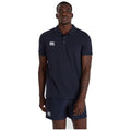 A man stands straight wearing a navy polo shirt and matching shorts with the logo of Canterbury visible on both garments against a plain white background.