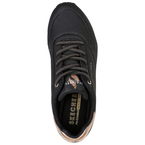 A black sneaker with perforated details and brown laces rests upright showcasing the Skechers logo and memory foam insole amidst a neutral background.