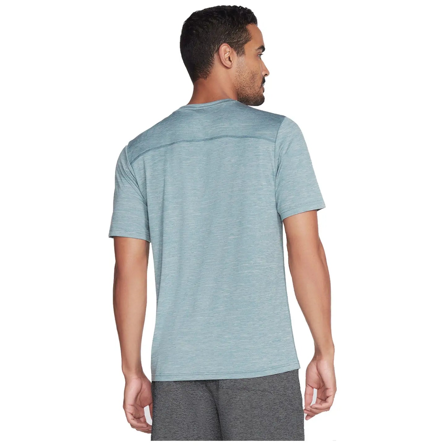 A man stands facing away wearing a light blue athletic shirt and gray shorts in a neutral setting likely for exercise or casual wear.