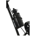 A black water bottle is mounted on a bike frame secured with a strap while the bike's wheel is partially visible in the lower right corner against a neutral background.