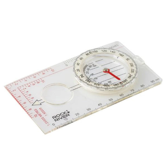 A clear plastic compass rests on a flat surface indicating directions with a red needle while surrounded by measurement markings for navigation purposes and a small magnifying lens.