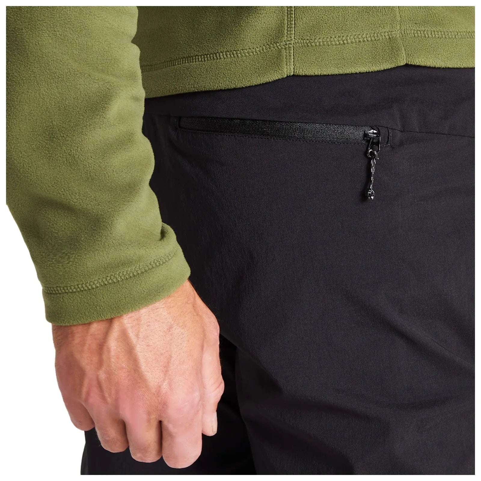 A close-up of a hand resting against a black pant pocket with a zipper while wearing a green fleece jacket in a neutral environment.