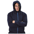 A man wearing a dark hooded jacket stands with arms crossed he appears serious and focused against a plain white background. The jacket features a quilted front and a logo on the sleeve.