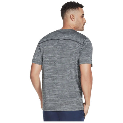 A man stands with his back facing the viewer wearing a short-sleeved, textured gray shirt and dark pants in a neutral background