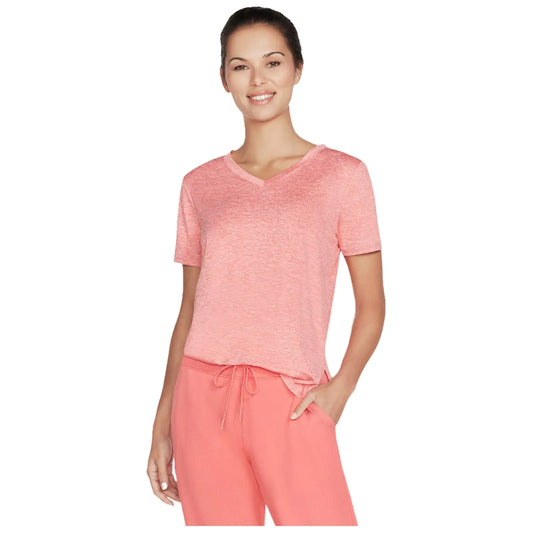 A woman stands wearing a pink v-neck t-shirt and matching drawstring pants smiling confidently while posing in a neutral background that emphasizes her casual style and comfort.