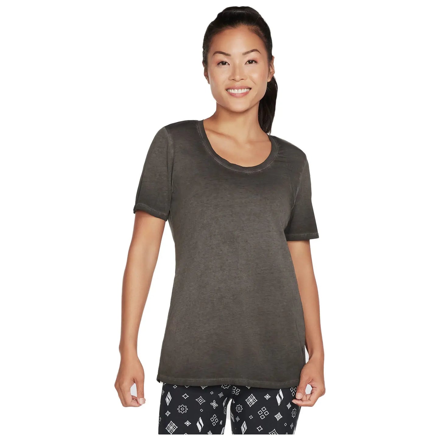 A woman smiles while wearing a dark shirt and patterned leggings standing in a neutral background that highlights her casual outfit and relaxed posture.