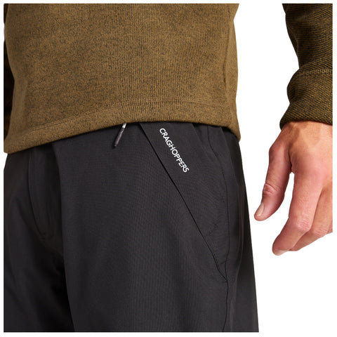 A close-up view of a person's torso wearing a brown sweater and black pants with a visible pocket featuring the logo Craghoppers, as a hand touches the fabric.