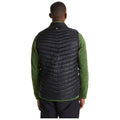 A person wearing a black puffy vest over a green long-sleeve shirt stands with their back facing the viewer in a neutral background, suggesting an outdoor or casual context.