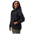 A woman wearing a black hoodie smiles while standing with her hand in the pocket of the hoodie against a plain white background. She has long hair and is wearing leggings.