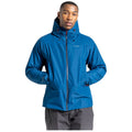 A man wears a blue jacket with a hood and zip closure while standing with his arms crossed in front of him against a plain white background.