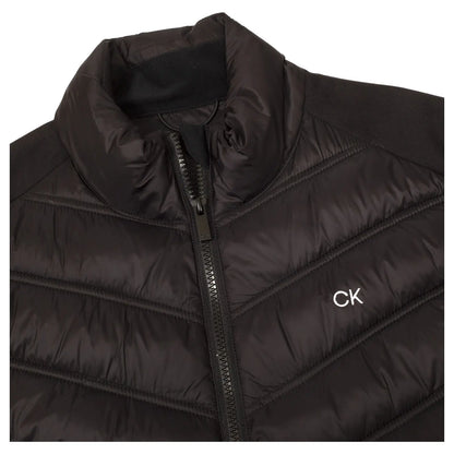 A black puffer jacket is laid flat displaying a high collar and a zippered front. The fabric has a quilted texture with a small logo embroidered on the chest.