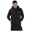 A man is standing with a slight smile wearing a black hooded sweatshirt with a logo on the chest and his hands in pockets against a plain white background.