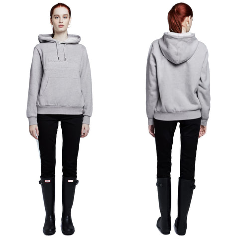 A person stands in a gray hoodie and black pants wearing tall black rubber boots showcasing a casual outfit in a minimalist setting with a white background.