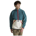 A person wearing a two-tone fleece pullover with a high collar stands, displaying a relaxed expression. The upper part is teal while the lower part is cream with red accents.