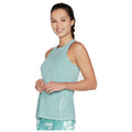 A woman stands smiling while wearing a light blue athletic tank top and patterned leggings her hands are clasped in front of her with a simple background